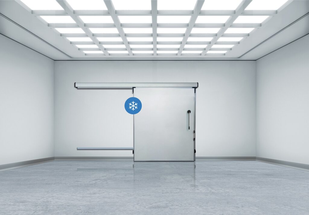 Industrial walk-in freezer sliding door under grid lights.