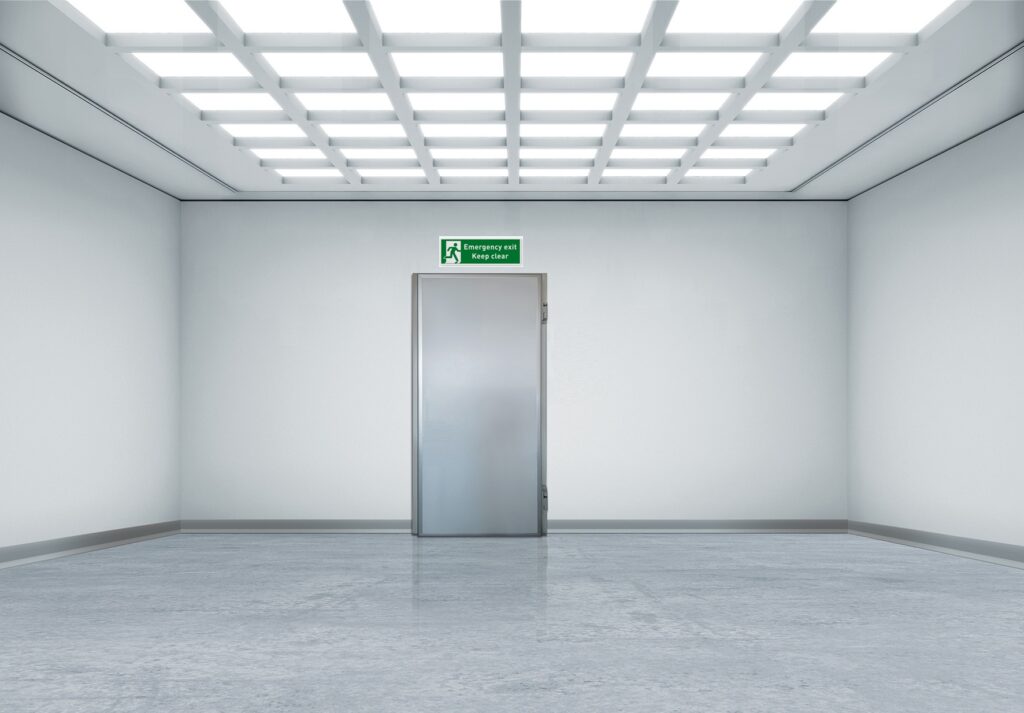 Empty white room with metal door and emergency exit sign above.